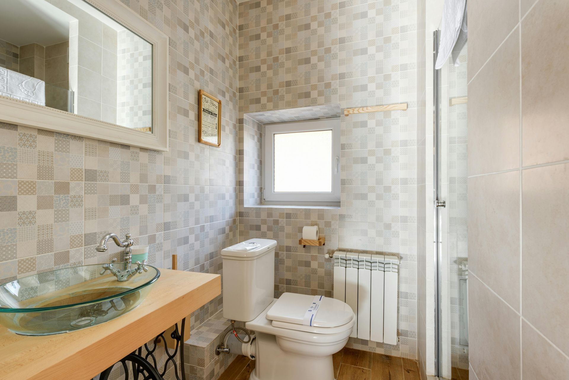 Small bathroom with mosaic tile walls, glass sink, toilet, window, and radiator.